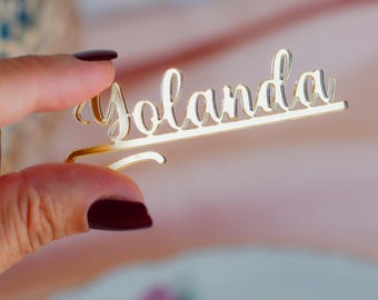 May include: A gold-colored, cursive nameplate that reads "Yolanda". The nameplate is held by a hand with dark red nail polish. The background is a soft pink and white.