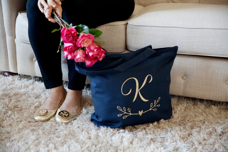 May include: A navy blue tote bag with a gold letter "K" and a small heart design. The bag is sitting on a light brown rug.