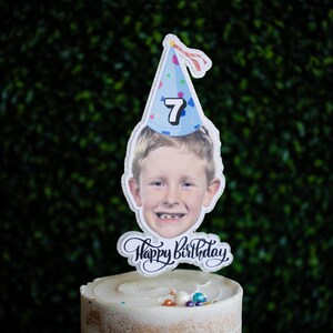 Może przedstawiać: Spersonalizowany topper na ciasto z fotografią dziecka w niebieskiej czapeczce urodzinowej z numerem 7. Słowa "Happy Birthday" są napisane kursywą pod zdjęciem. Topper umieszczony jest na lukrowanym torcie.