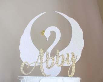 May include: White glitter swan cake topper with gold glitter wings and a gold star on its head. The name "Abby" is written in gold glitter below the swan.