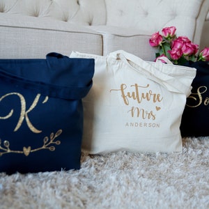 May include: Three tote bags with gold lettering. The first bag is navy blue with a gold letter 'K' and a gold heart with branches on either side. The second bag is white with the text 'future Mrs. ANDERSON' in gold. The third bag is black with the name 'Sophia' in gold.