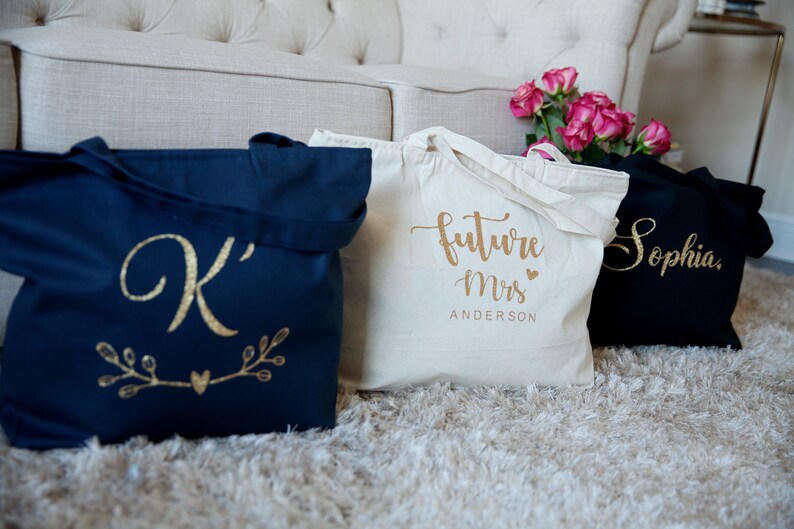 May include: Three tote bags with gold lettering. The first bag is navy blue with a gold letter 'K' and a gold heart with branches on either side. The second bag is white with the text 'future Mrs. ANDERSON' in gold. The third bag is black with the name 'Sophia' in gold.