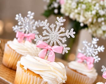 May include: Cupcakes topped with silver glitter snowflake decorations, each featuring a small light pink satin ribbon bow tied around the base of the snowflake topper. The cupcakes have white frosting and light-colored paper liners.