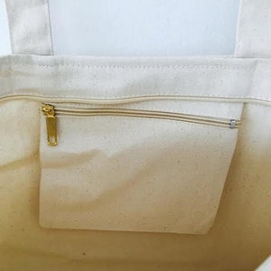 May include: A white canvas tote bag with a zippered pocket on the inside. The zipper is gold metal.