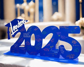 May include: A blue graduation sign with the year 2025 cut out. The sign includes a graduation cap with the name "Landon" and is covered in handwritten messages. The sign is on a white surface.