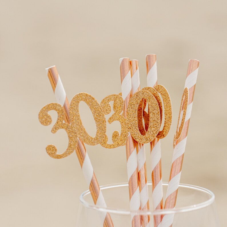 May include: Gold glitter straws with the number "30" on them. The straws are striped with white and rose gold.