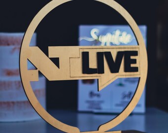 May include: A wooden sign with the text "NT LIVE" in black letters. The sign is in a circular frame with a rectangular base. The background is blurred, with a hint of a sign that says "Synaptic".