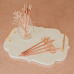 May include: A white marble tray with gold handles, featuring rose gold straws with glitter numbers "30" and a glass with rose gold straws.