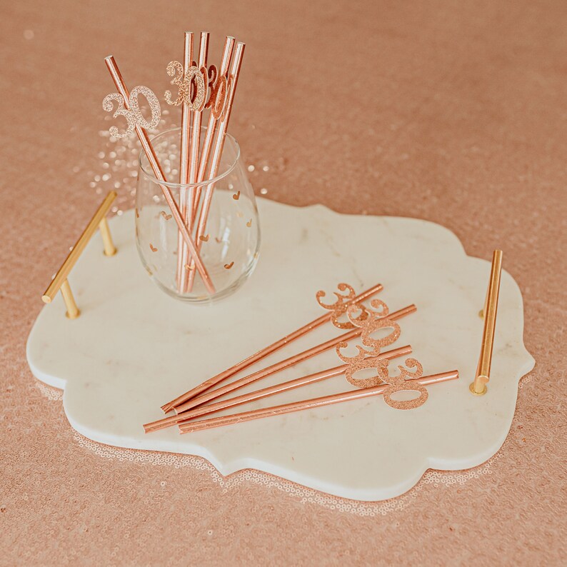 May include: A white marble tray with gold handles, featuring rose gold straws with glitter numbers "30" and a glass with rose gold straws.