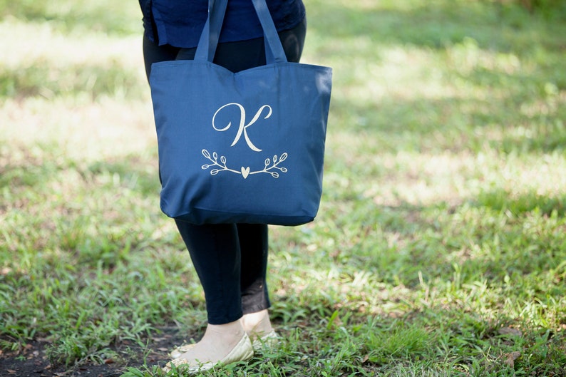 May include: A navy blue tote bag with a white monogram of the letter "K" and a small heart with branches on either side.