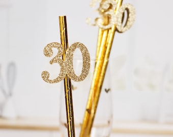 May include: Gold glitter straws with the number "30" on them. The straws are in a clear glass with a gold rim.