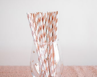 May include: A clear glass vase filled with a bundle of white and rose gold striped paper straws.