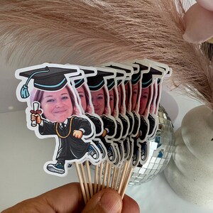 May include: A set of graduation cap toppers with personalised photos. Each topper features a cartoon graduate wearing a cap and gown, holding a diploma. The toppers are attached to wooden skewers.