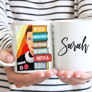 May include: Two white ceramic mugs. One mug features a colorful illustration of Ruth Bader Ginsburg with the text "Never underestimate the power of a girl with a book." The other mug has the name "Sarah" written in black cursive.
