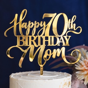 May include: A gold-colored acrylic cake topper featuring the text Happy 70th BIRTHDAY Mom in a combination of cursive and block lettering. The topper has a reflective, metallic finish and includes a single vertical stake at the base.