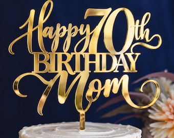 May include: A gold-colored acrylic cake topper featuring the text Happy 70th BIRTHDAY Mom in a combination of cursive and block lettering. The topper has a reflective, metallic finish and includes a single vertical stake at the base.