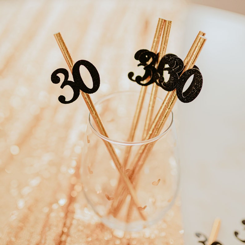 May include: Gold straws with black glitter numbers 30 attached to them. The straws are in a clear glass.