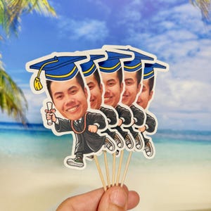 May include: Set of five graduation cupcake toppers featuring cartoon illustrations of a person wearing a blue graduation cap and gown, holding a diploma. The toppers are printed on white cardstock and attached to wooden skewers.