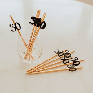 May include: Gold straws with black glitter 30s attached. The straws are in a clear glass with a heart pattern.