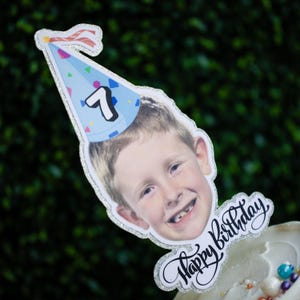 Może przedstawiać: Topper na tort urodzinowy przedstawiający uśmiechnięte dziecko w niebieskiej czapeczce z numerem 7. Słowa "Happy Birthday" są napisane kursywą pod twarzą dziecka. Krawędzie są obrysowane brokatem.