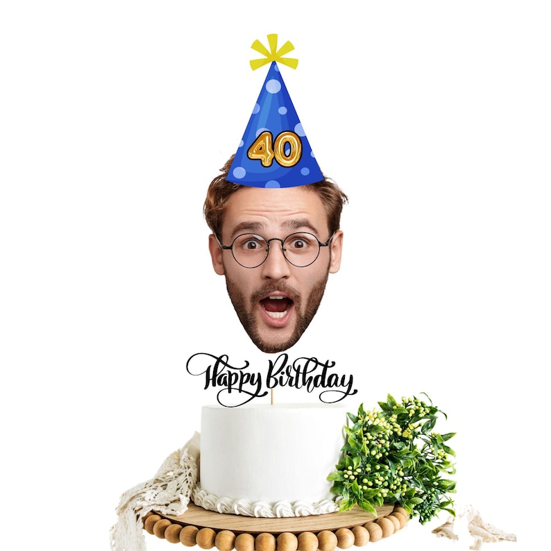 Może przedstawiać: Białe ciasto urodzinowe z topperem "Happy Birthday", zieloną rośliną i drewnianym stojakiem na ciasto. Niebieska czapeczka urodzinowa z numerem 40 znajduje się nad ciastem, z twarzą mężczyzny w okularach.
