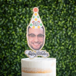 Może przedstawiać: Spersonalizowany topper na tort urodzinowy z uśmiechniętą osobą w okularach i czapeczce urodzinowej z numerem 30. Topper ma napis "Happy Birthday" i jest umieszczony na białym torcie.