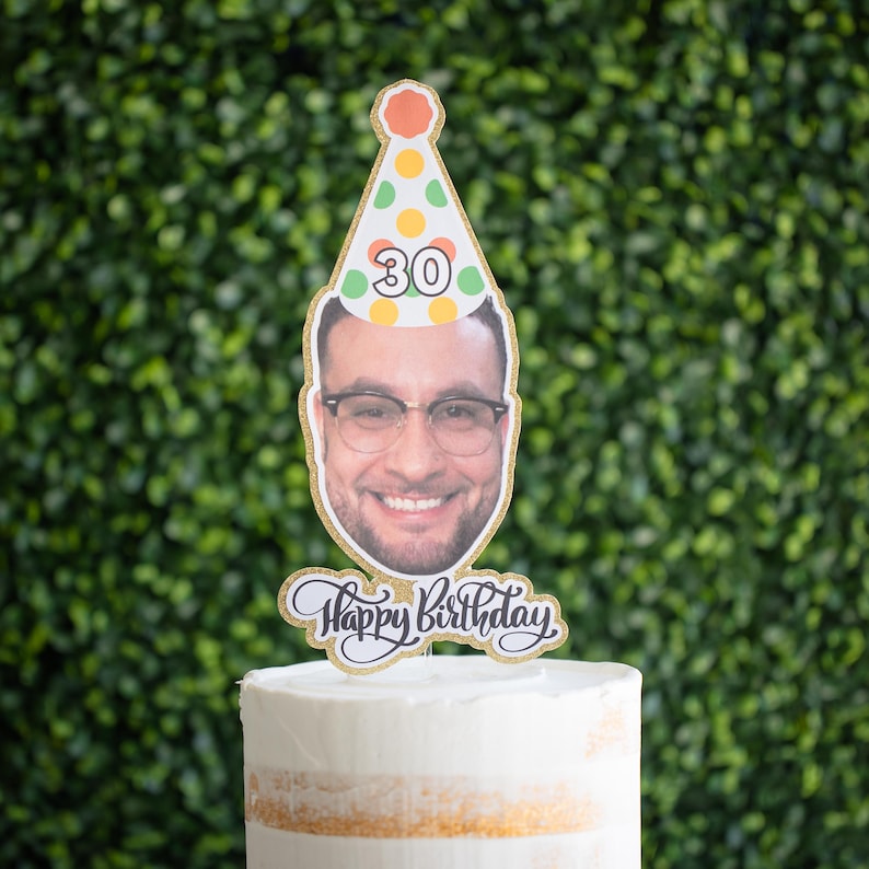 Może przedstawiać: Spersonalizowany topper na tort urodzinowy z uśmiechniętą osobą w okularach i czapeczce urodzinowej z numerem 30. Topper ma napis "Happy Birthday" i jest umieszczony na białym torcie.