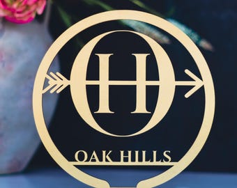 May include: A gold-colored wooden sign with the text "OAK HILLS" at the bottom. The sign features a circular design with the letters "OH" in the center, intersected by an arrow. The sign is mounted on a small wooden base.