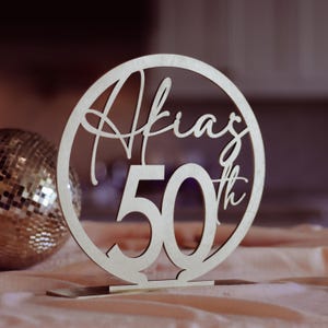 May include: A wooden table decoration with the name "Akia" and the number "50th" cut out in a circular design. The decoration is made of light-colored wood and stands on a small base. A disco ball is visible in the background.