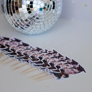 May include: A row of graduation themed cupcake toppers with a person's face on each one. The toppers are on wooden skewers and are arranged in a row on a white surface.