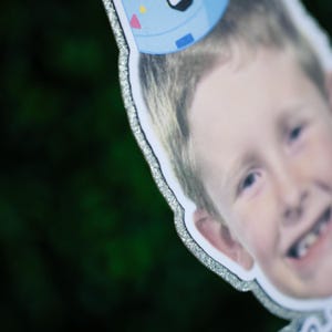 Może przedstawiać: Wycięcie z kartonu uśmiechniętego dziecka w niebieskiej czapeczce urodzinowej z kolorowymi akcentami. Wycięcie ma srebrną, błyszczącą obw&oacute;dkę na rozmytym zielonym tle.