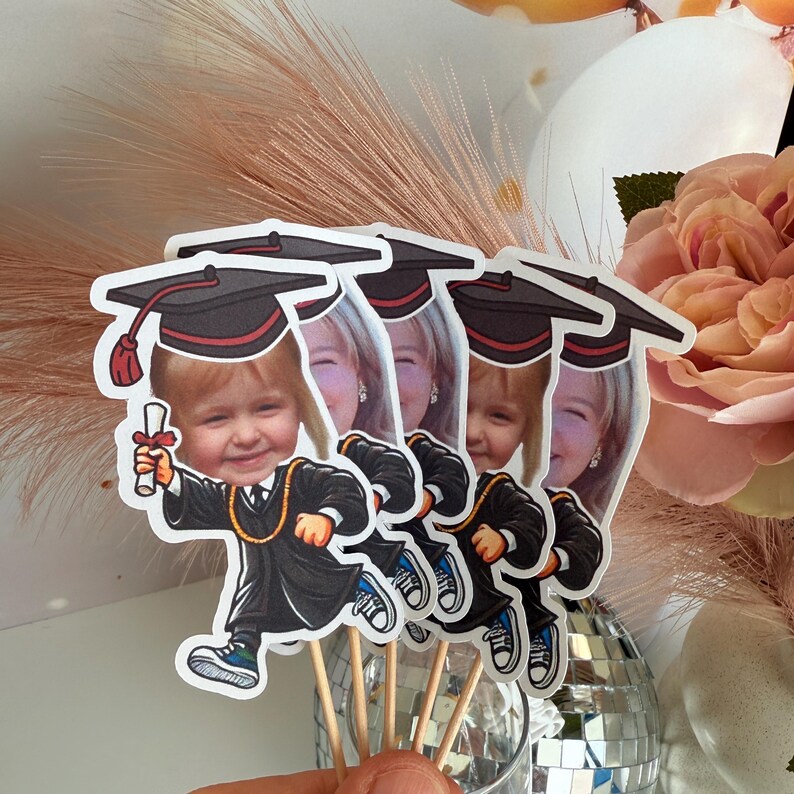 May include: Four graduation cap toppers featuring photographs of children. Each topper is a cartoon character wearing a black graduation cap and gown. The toppers are on wooden sticks.
