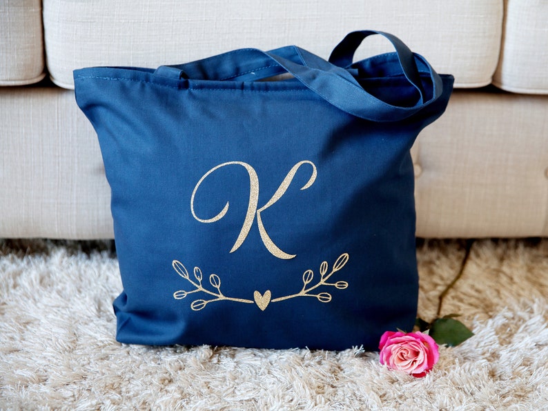 May include: A navy blue tote bag with a gold glitter letter "K" and a heart design. The bag has two handles and is made of canvas.