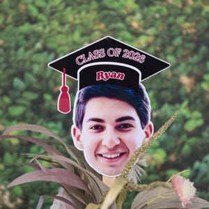 May include: A graduation photo prop featuring a smiling young person's face with a graduation cap. The cap is black with the text "CLASS OF 2026" and the name "Ryan". The prop is held up by a wooden stick.