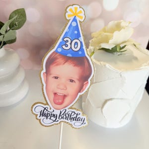 Może przedstawiać: Spersonalizowany topper na tort ze zdjęciem osoby w niebieskiej czapeczce urodzinowej z numerem 30. Topper ma złotą, błyszczącą obw&oacute;dkę i napis "Happy Birthday" w ozdobnej czcionce.