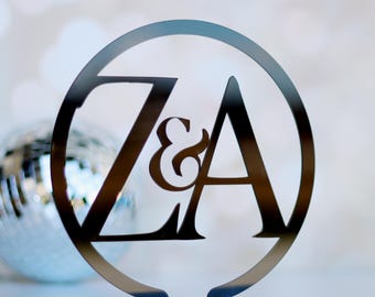 May include: A metallic silver wedding cake topper featuring the letters "Z & A" within a circular frame. The letters are connected by an ampersand. The topper is mounted on a rectangular base, with a disco ball in the background.