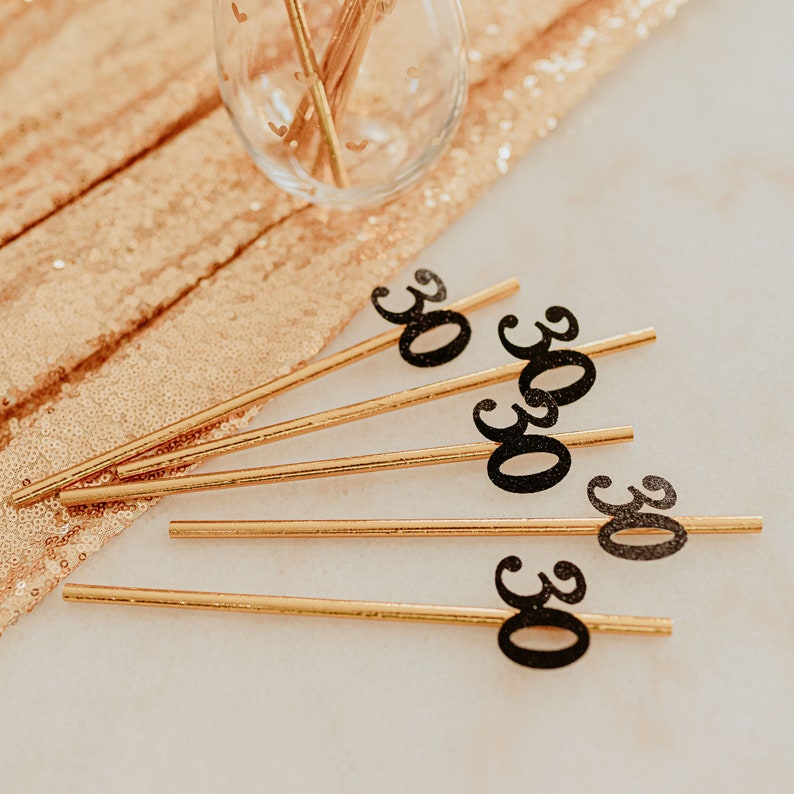May include: Five gold straws with black glitter 30s on them. The straws are on a white surface with a gold sequin tablecloth in the background.