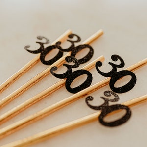 May include: Five gold straws with black glitter numbers "30" on them. The straws are arranged in a fan shape.
