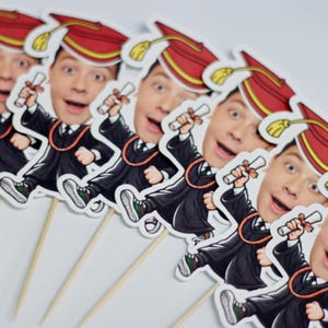 May include: Set of six graduation cupcake toppers featuring a cartoon graduate wearing a cap and gown, holding a diploma, and a red and yellow tassel. Each topper is attached to a wooden stick.