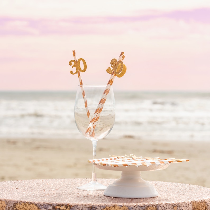 May include: Two gold glitter straws with the number "30" on them are in a clear wine glass. The glass is on a white cake stand with a gold glitter paper doily. The background is a blurry image of a beach with a pink and orange sunset.