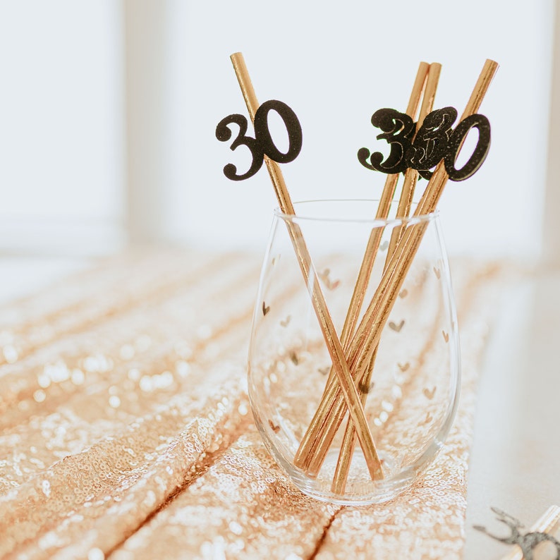 May include: Four gold straws with black paper cutouts of the number "30" attached to them. The straws are in a clear glass with a gold heart pattern. The glass is sitting on a gold sequin tablecloth.