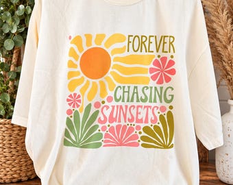 May include: A cream-colored t-shirt featuring a graphic print of a yellow sun, pink flowers, and green foliage. The text "FOREVER CHASING SUNSETS" is printed in a mix of green and pink lettering across the center of the design.