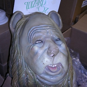 May include: A green and white cardboard box with the text "The Wizard of Oz" on it. A gray and brown mask of the Wicked Witch of the West from the Wizard of Oz movie is on top of the box. The mask has a wide, open mouth and a long, flowing wig.