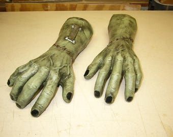 Può includere: Un paio di mani verdi, simili a quelle di uno zombie, con unghie nere. Le mani hanno un aspetto cucito ai polsi e dettagli metallici. Probabilmente parte di un costume o di un accessorio di Halloween.