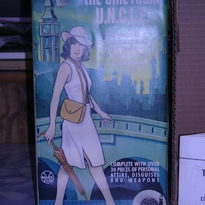 May include: A vintage box for a dollhouse featuring a woman in a white dress and a hat, holding an umbrella. The box has a blue and green background with the text "The Girl From U.N.C.L.E." and "Complete with over 30 pieces of personal attire, disguises and weapons".