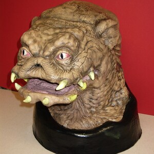 May include: A detailed, hand-painted, brown and gray creature bust with a toothy grin and large, yellow eyes. The bust is mounted on a black base.