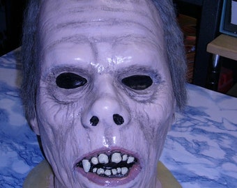 Può includere: Maschera dettagliata di un volto invecchiato con pelle pallida, orbite scure e una bocca spalancata che rivela denti storti. La maschera ha i capelli grigi ed è probabilmente per Halloween o costumi.