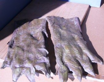 May include: Two green and brown textured fabric monster hands with claws. The hands are laid out flat on a wooden surface.