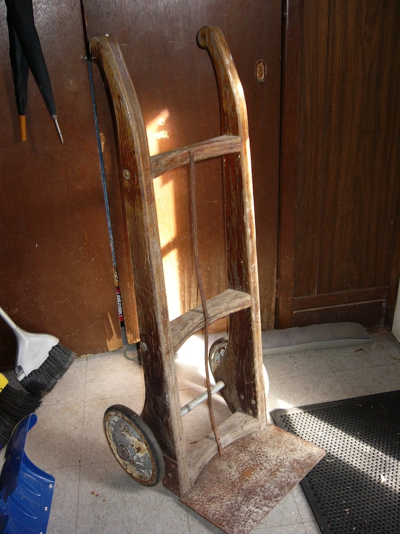 May include: A wooden hand truck with a metal handle and two wheels. The truck is made of weathered wood and has a rusty metal base.
