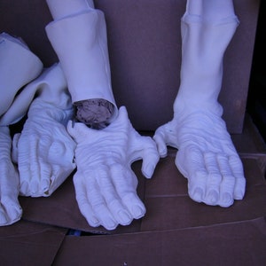 May include: Several off-white, textured prosthetic limbs, including feet, hands, and lower legs, are arranged inside a cardboard box. The pieces have detailed skin-like textures and are likely for costume or special effects use.